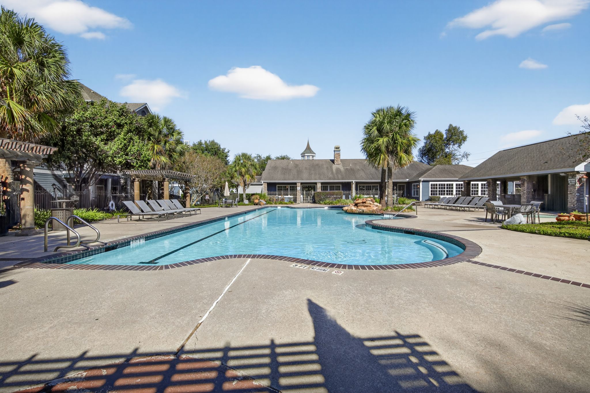 Spacious community pool with curved design, sun loungers, shaded seating areas, and clubhouse buildings framed by palm trees
