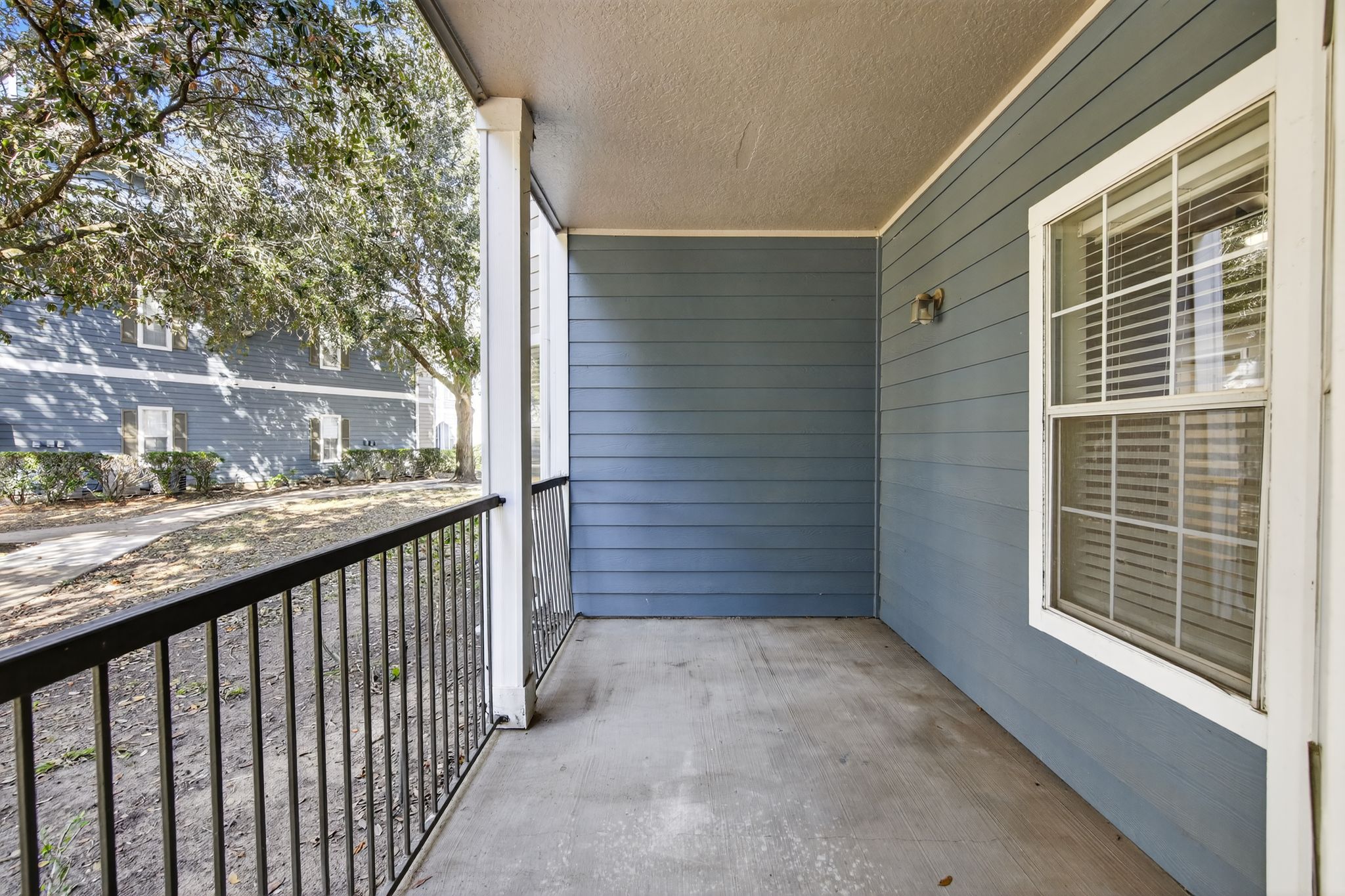Covered patio with blue siding, black railing, and a view of nearby buildings and trees in the community