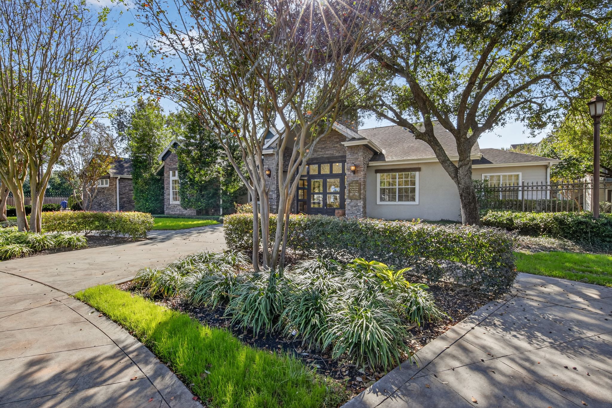 Landscaped clubhouse approach with mature trees, trimmed shrubs, stone details, and paved walkways under clear skies