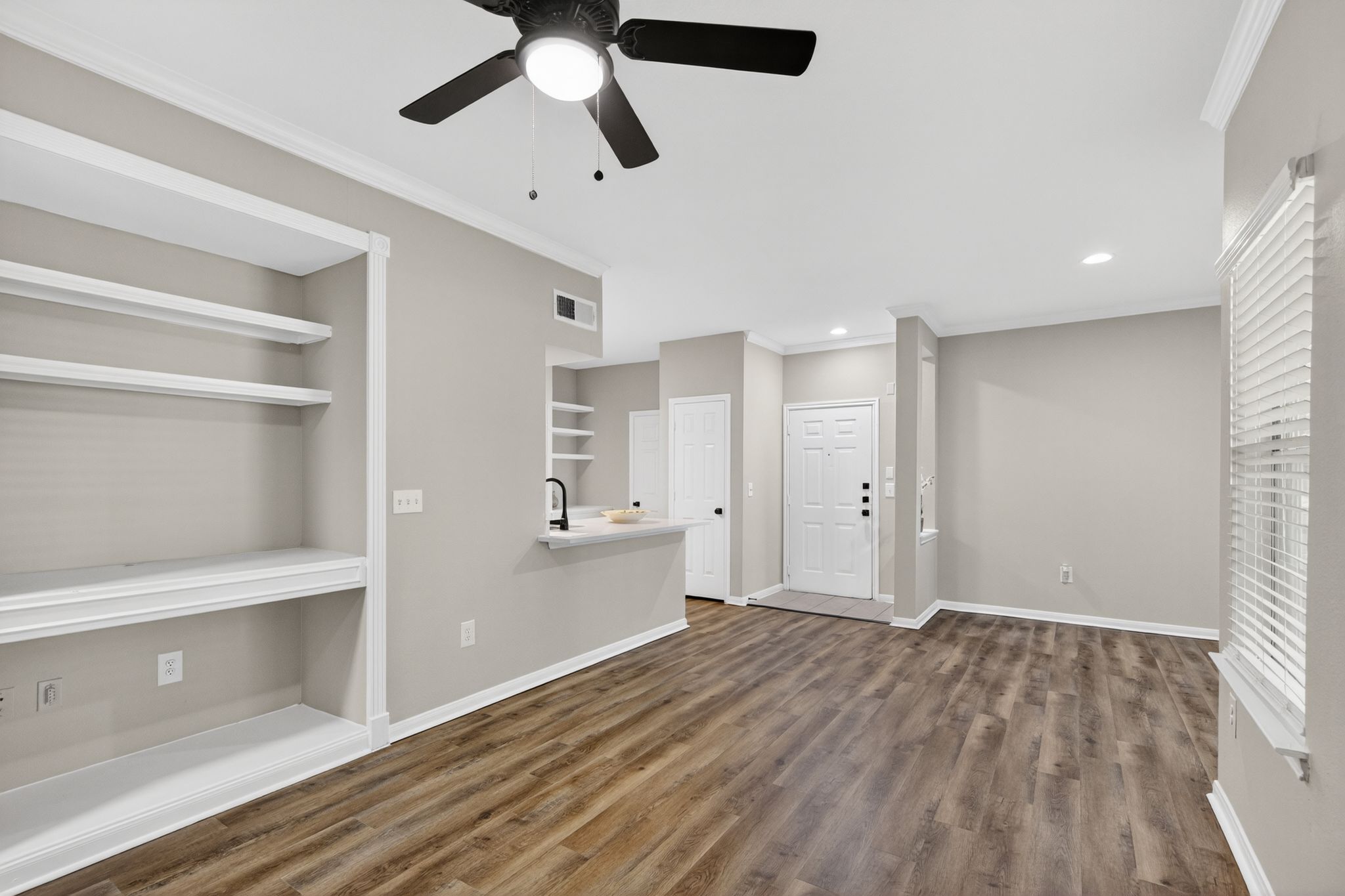 Open living space with wood style flooring, built in shelving, ceiling fan, and a view into the kitchen and entryway