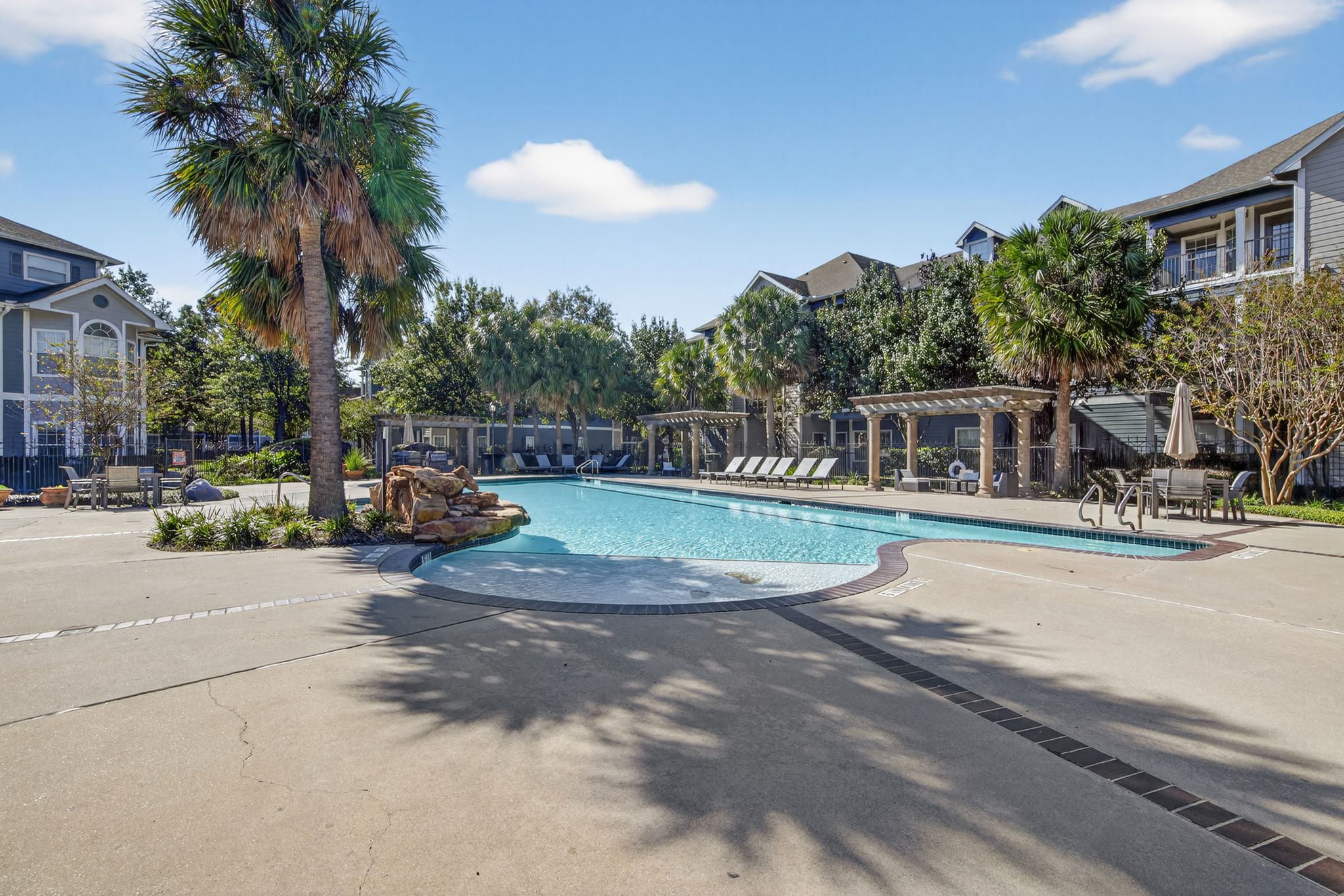 Resort-style swimming pool with palm trees, pergolas, lounge seating, and surrounding apartment buildings under a bright blue sky