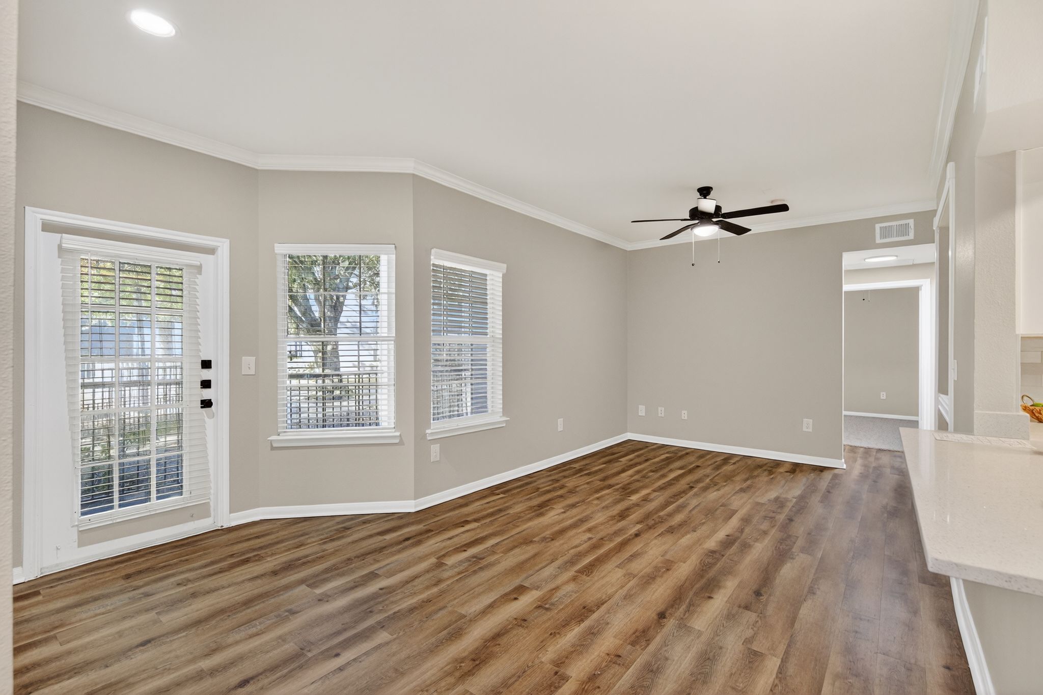 Spacious living area with wood style flooring, large windows, a ceiling fan, and a door leading to the outdoor patio