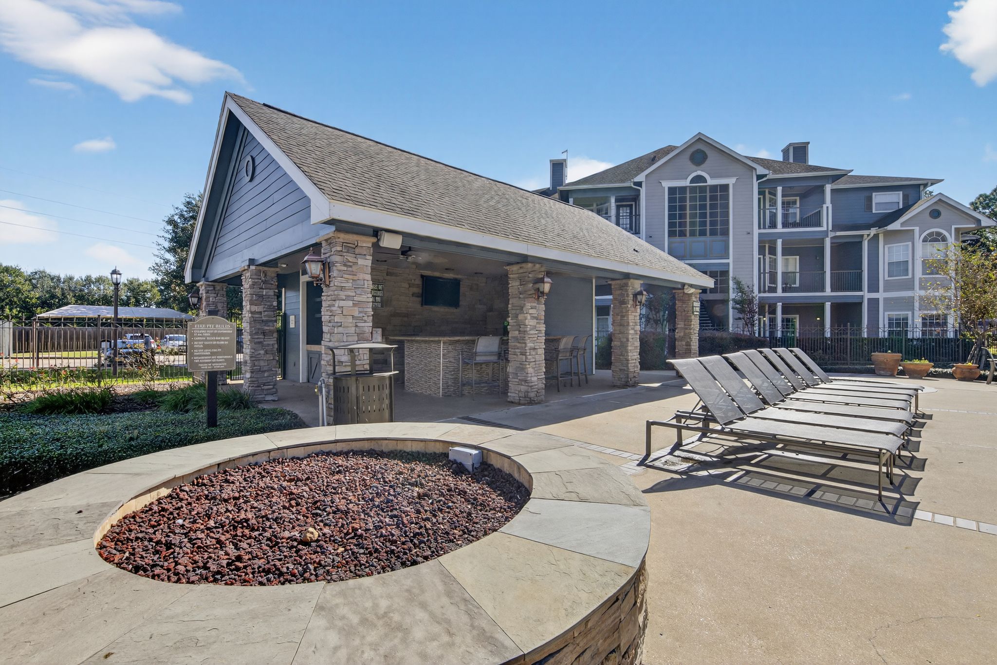Pool deck area with sun loungers, covered pavilion bar, stone fire pit feature, and apartment buildings in the background