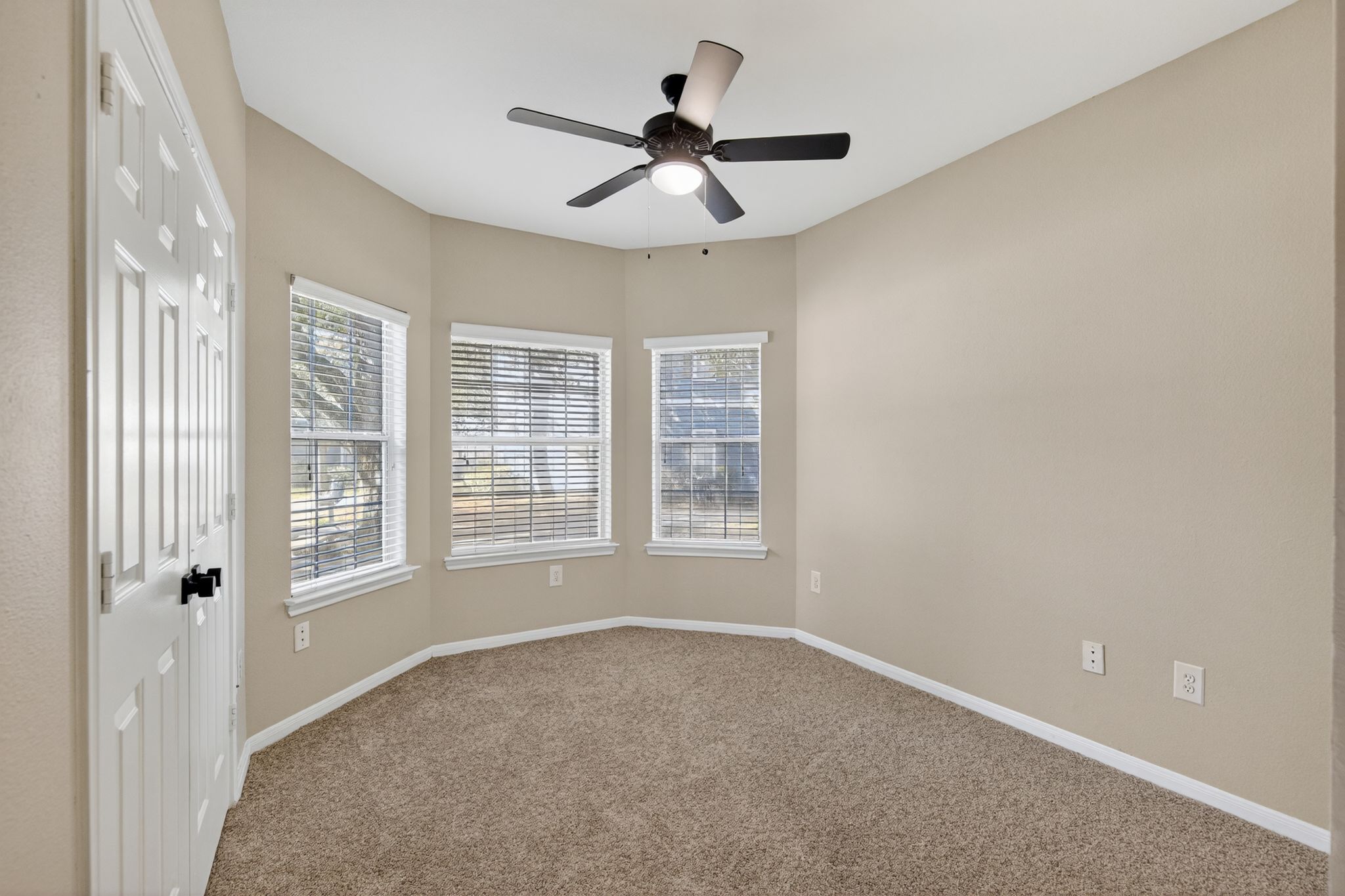 Empty bedroom with bay windows, neutral walls, carpeted flooring, and a ceiling fan centered in the space