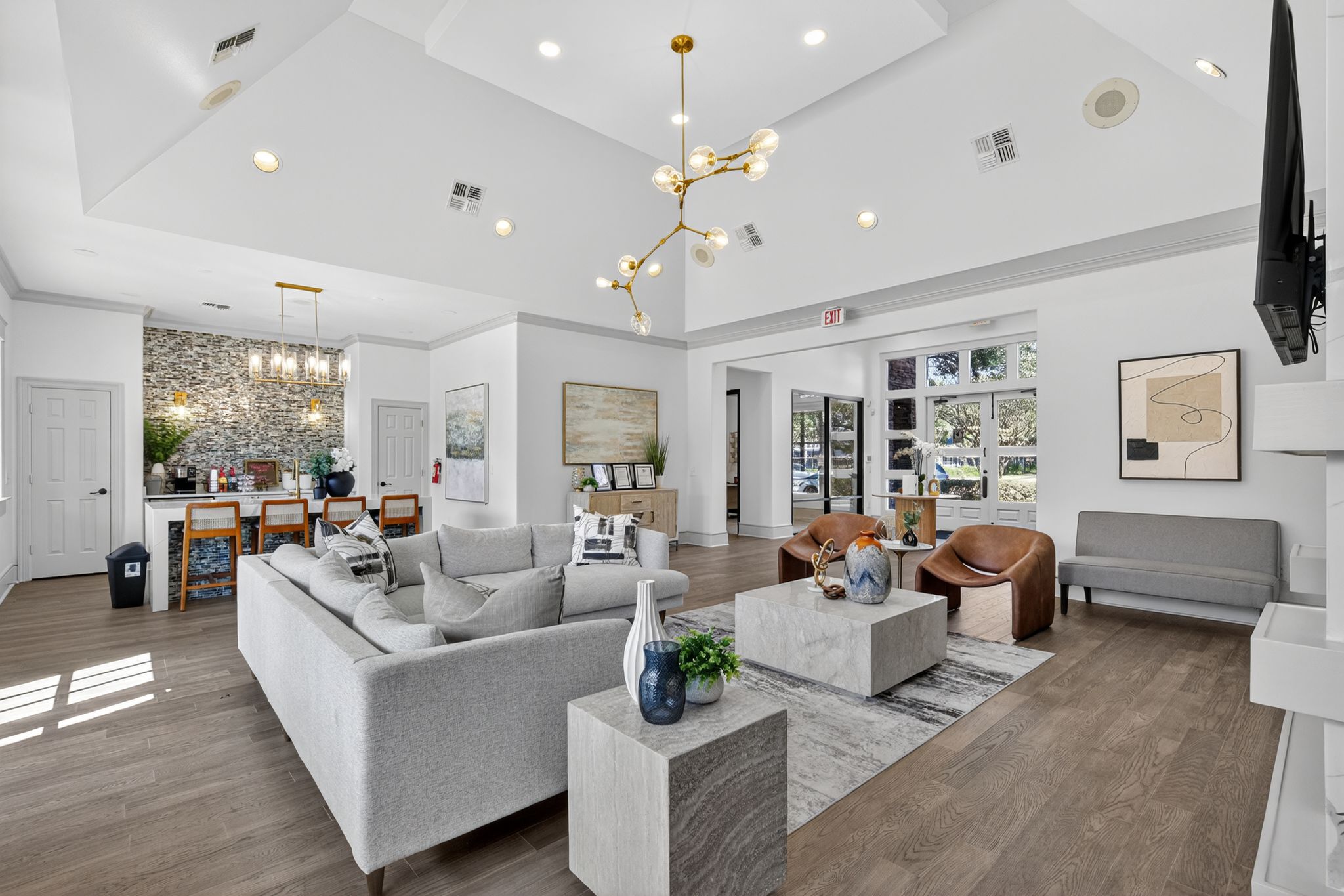 Bright resident lounge with modern sofas, accent chairs, statement lighting, wood floors, and an open refreshment bar area