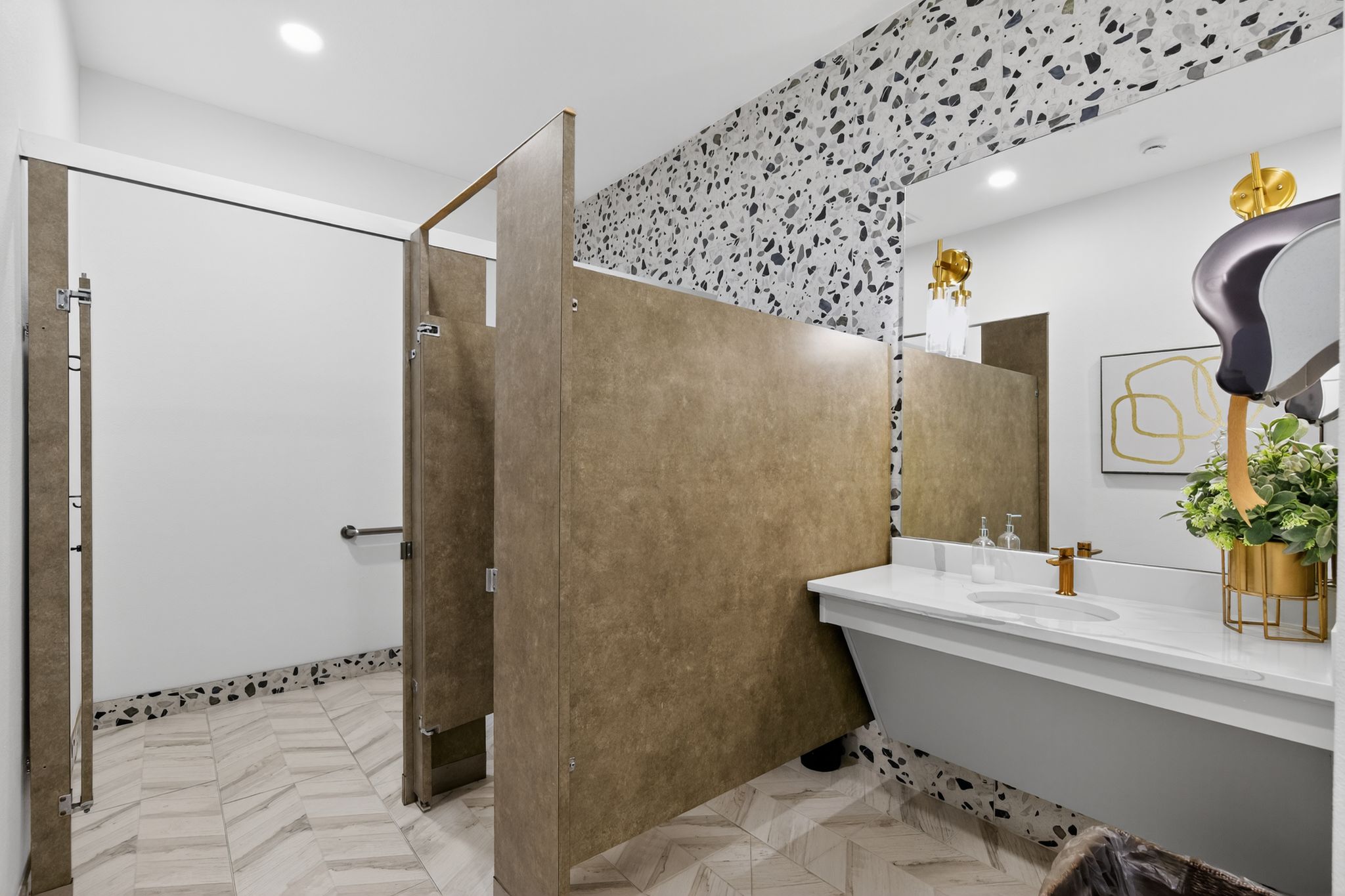 Updated community restroom with modern vanity, gold fixtures, privacy partitions, patterned tile flooring, and contemporary wall accents