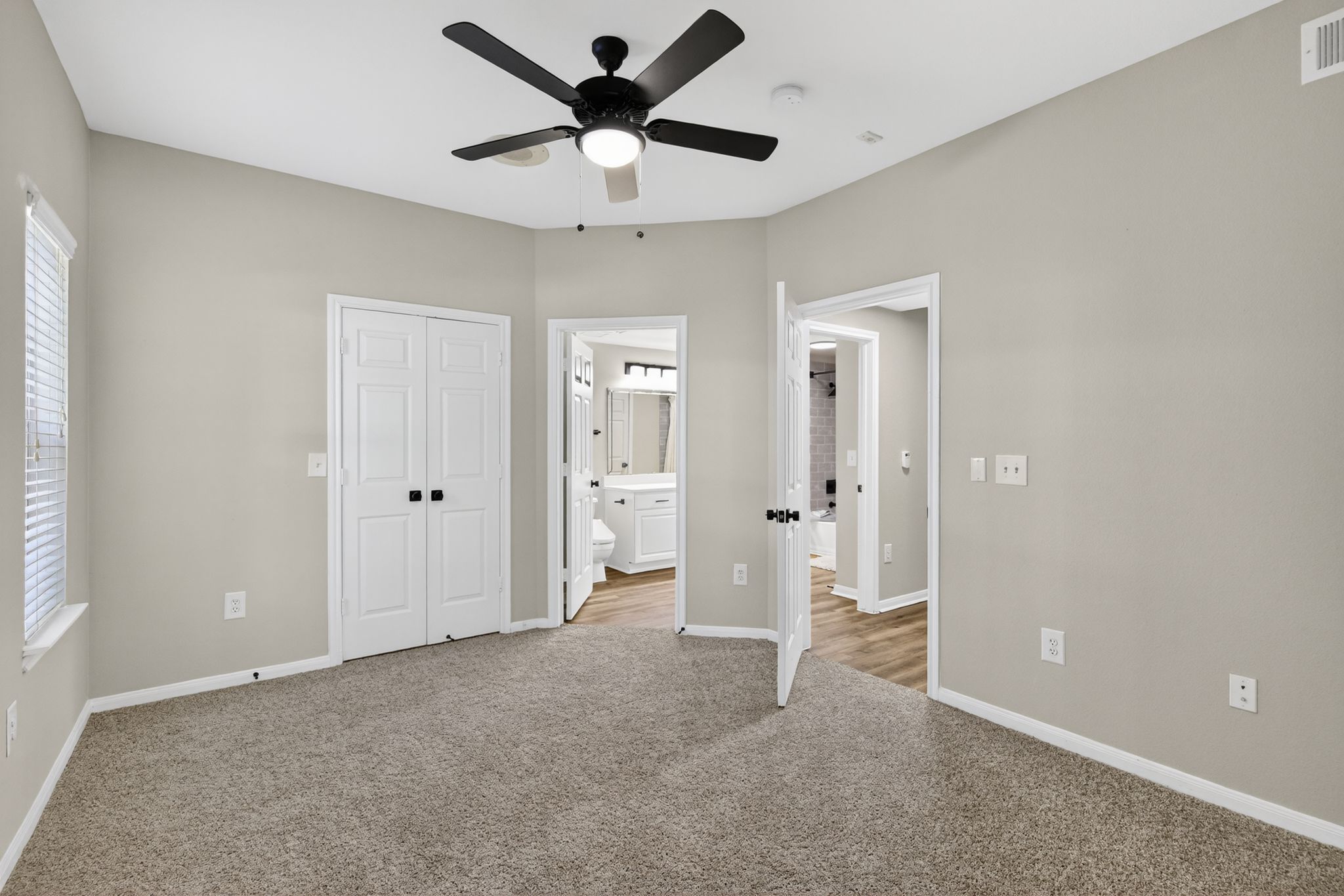 Bedroom with carpeted flooring, white double doors, ceiling fan, and open entrances leading to the bathroom and hallway