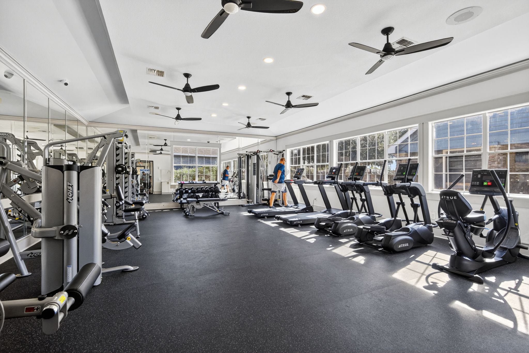 Fully equipped fitness center with treadmills, strength machines, free weights, ceiling fans, and wall mirrors in a bright space