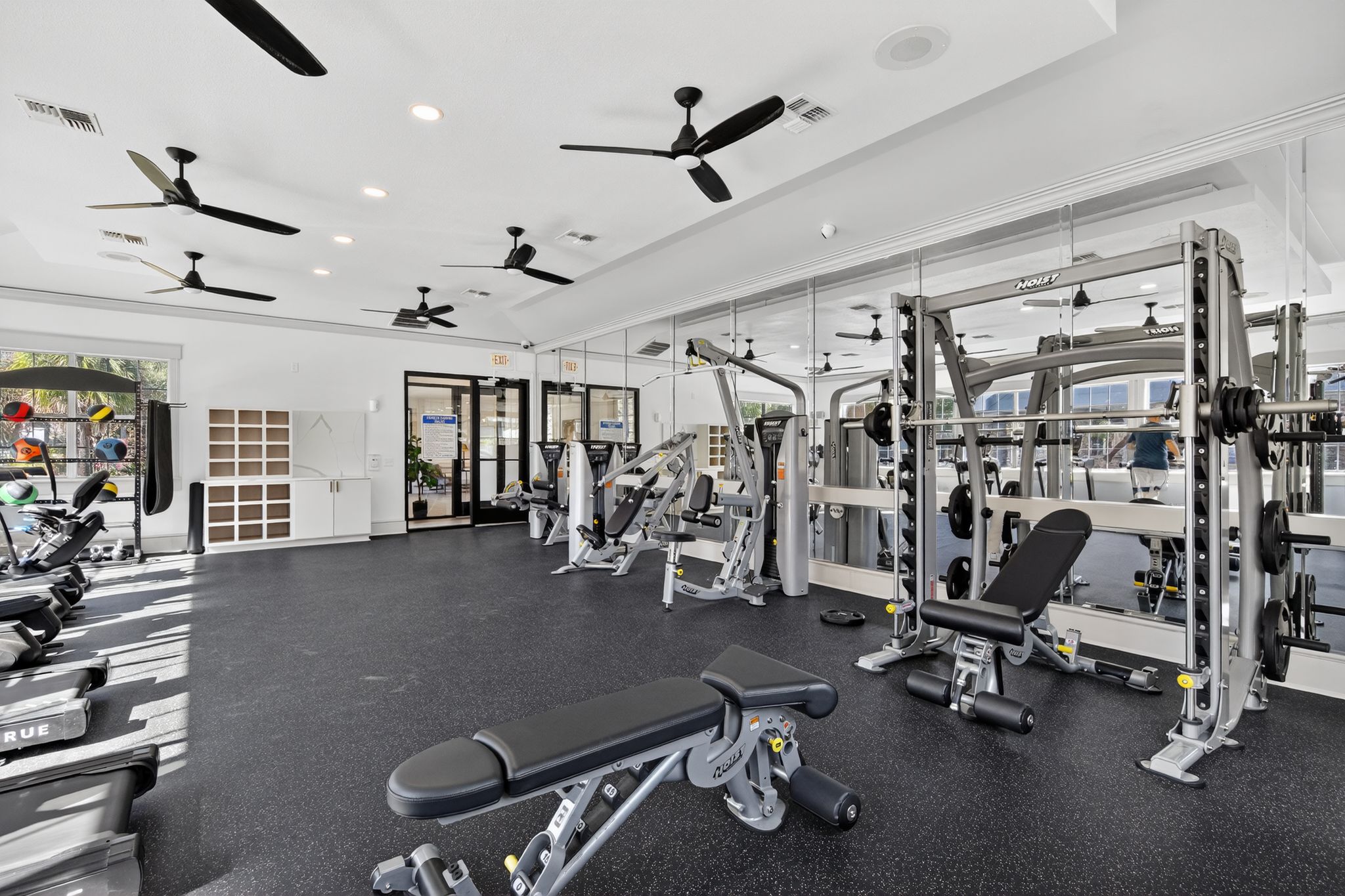 Modern apartment gym showcasing weight machines, benches, mirrored walls, rubber flooring, and ample natural light from windows