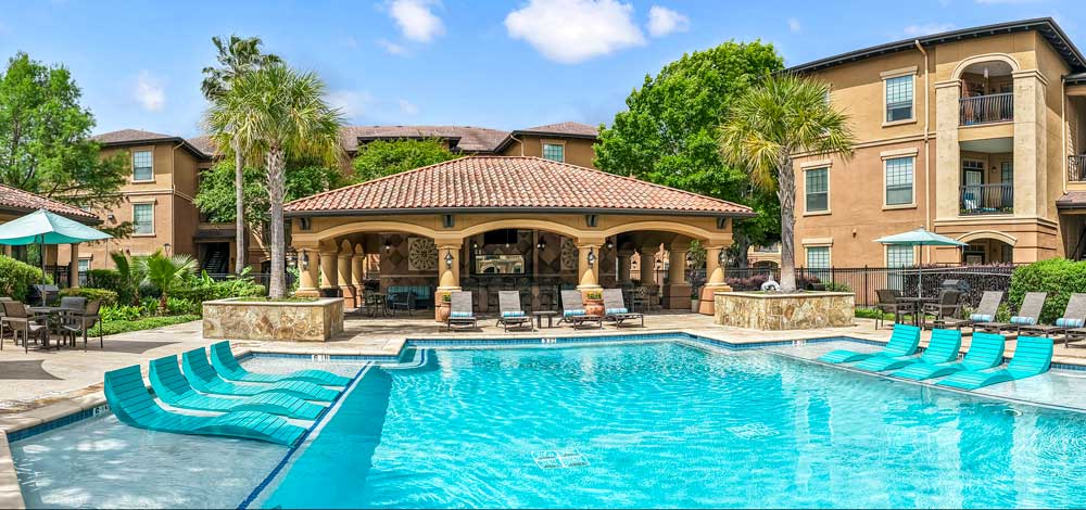 Pool with in-water loungers, pavilion with columns, palm trees, and surrounding apartment buildings in courtyard setting
