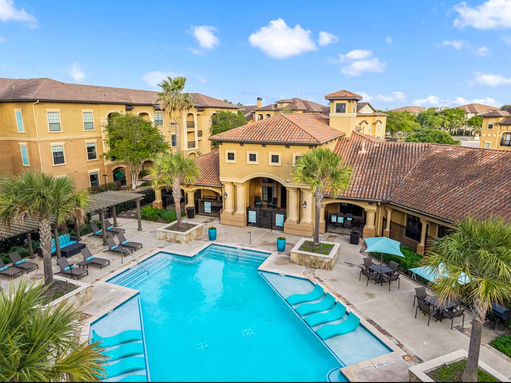 Aerial view of pool courtyard with in-water loungers, pergola seating, palm trees, and surrounding apartment buildings