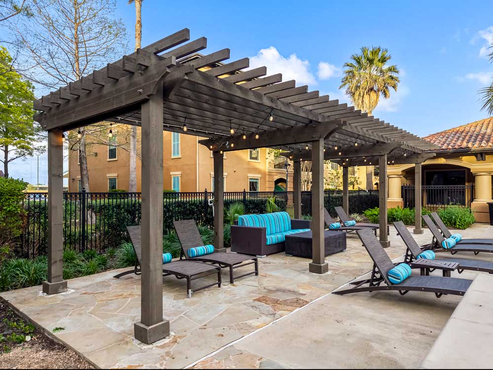 Pergola with string lights, cushioned seating, and lounge chairs beside landscaped courtyard and fenced apartment grounds