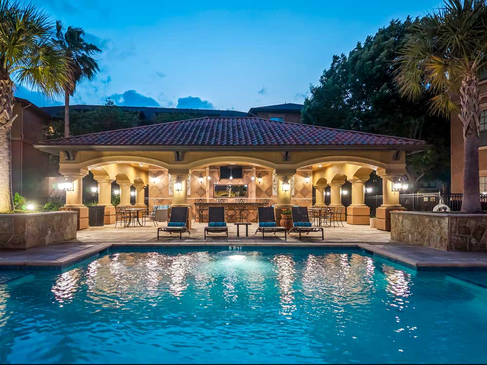 Evening pool view with illuminated pavilion, palm trees, and lounge seating reflecting across calm water surface