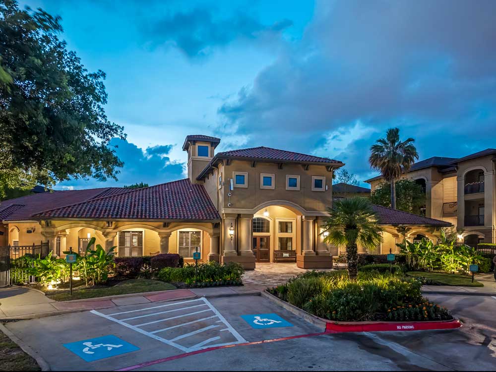 Clubhouse exterior at dusk with arched entry, illuminated windows, landscaped gardens, and accessible parking spaces nearby