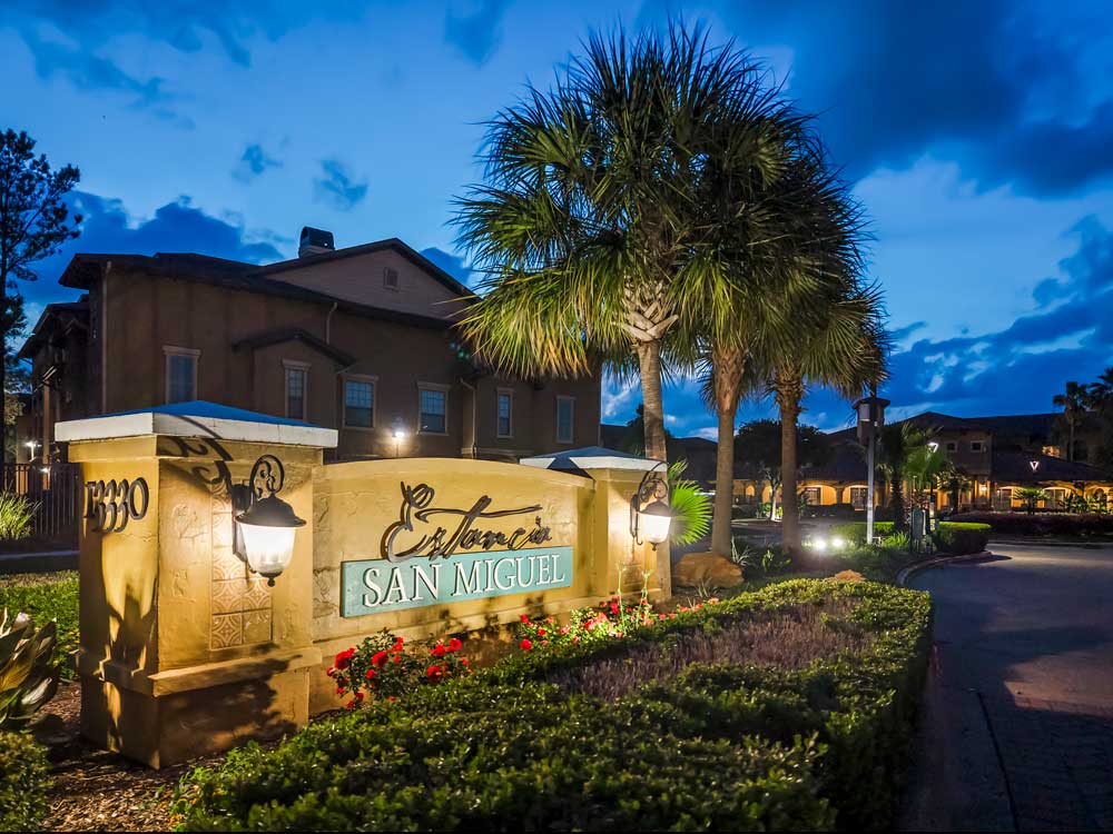 Community entrance sign with Estancia San Miguel lettering, decorative lamps, landscaped shrubs, and palm trees at dusk