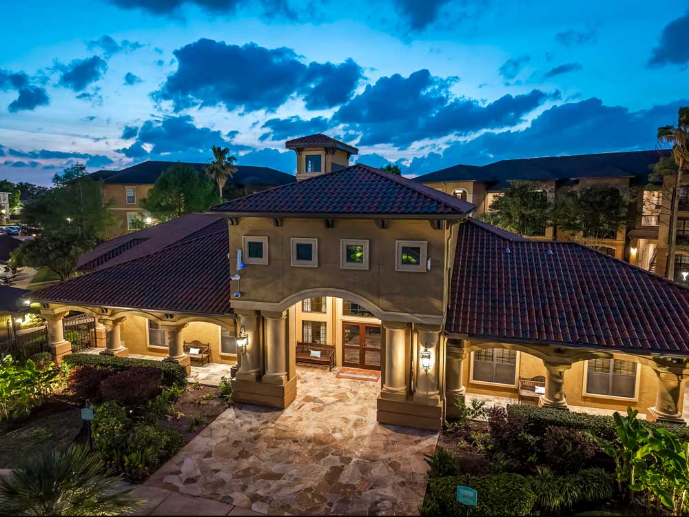 Elevated view of clubhouse with tiled roof, arched entry, landscaped grounds, and surrounding apartment buildings at sunset
