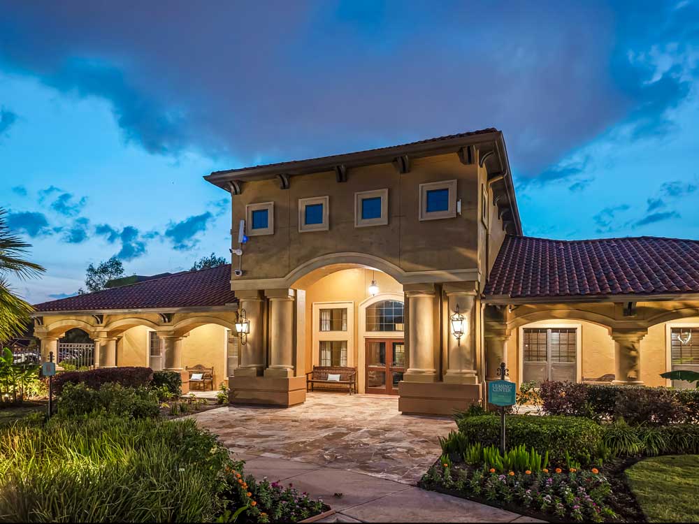 Leasing center exterior with arched entrance, warm lighting, landscaped walkways, and surrounding plants at dusk