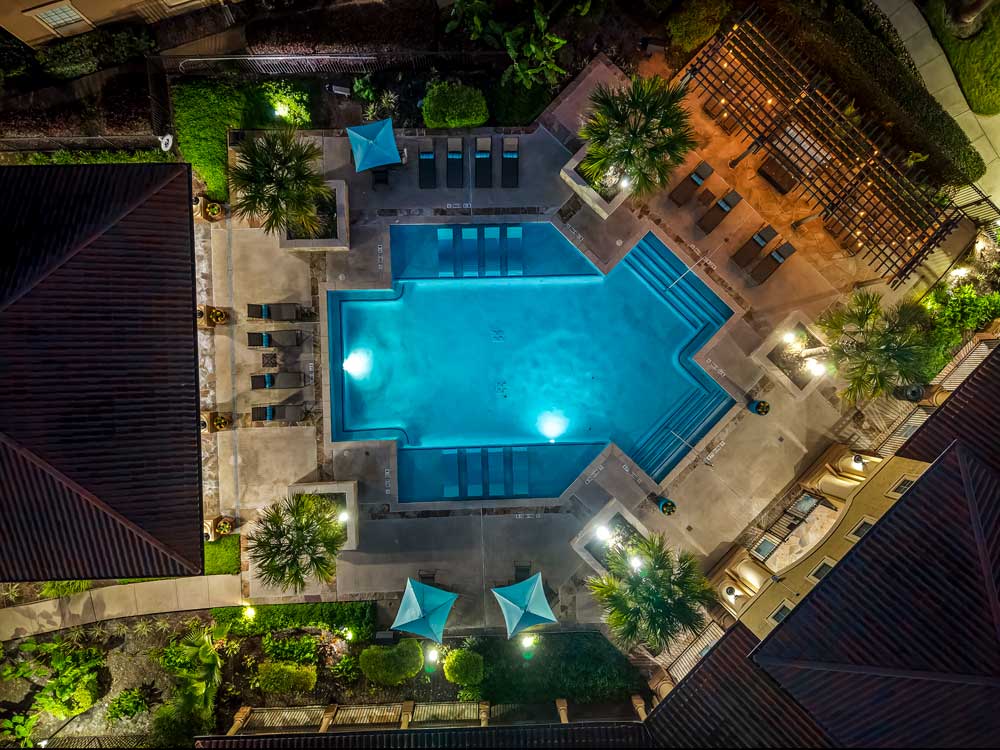 Nighttime aerial view of an illuminated pool deck with lounge seating, teal shade sails, palm trees, and a lit pergola