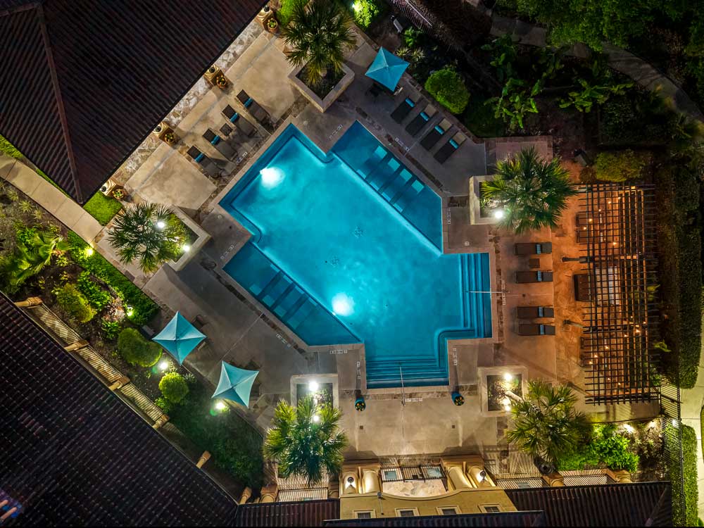 Aerial night view of a resort-style pool surrounded by lounge chairs, teal umbrellas, lush landscaping, and a pergola