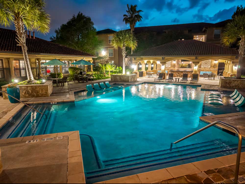 Nighttime pool with glowing water, palm trees, lit pavilion, and outdoor seating areas creating warm evening atmosphere
