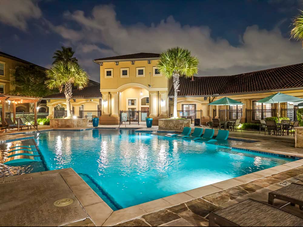Night view of resort-style pool with in-water loungers, palm trees, lit clubhouse, and outdoor seating areas