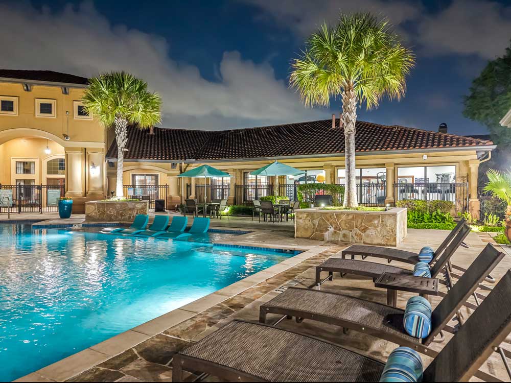 Evening poolside with lounge chairs, palm trees, lit clubhouse, and shaded seating areas overlooking illuminated water