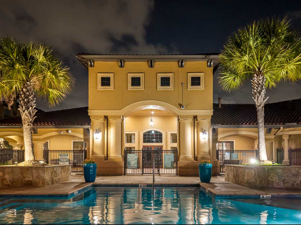 Symmetrical view of pool and clubhouse entrance framed by palm trees, outdoor lighting, and gated access in apartment community