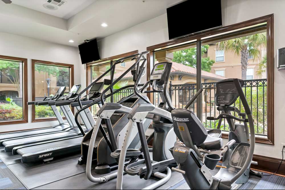 Fitness center with treadmills, elliptical machines, mounted TVs, and large windows overlooking landscaped courtyard area