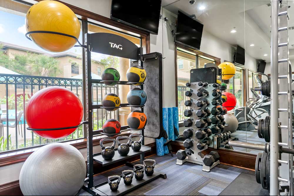 Fitness center with medicine balls, kettlebells, and dumbbells near large windows overlooking pool and landscaped courtyard