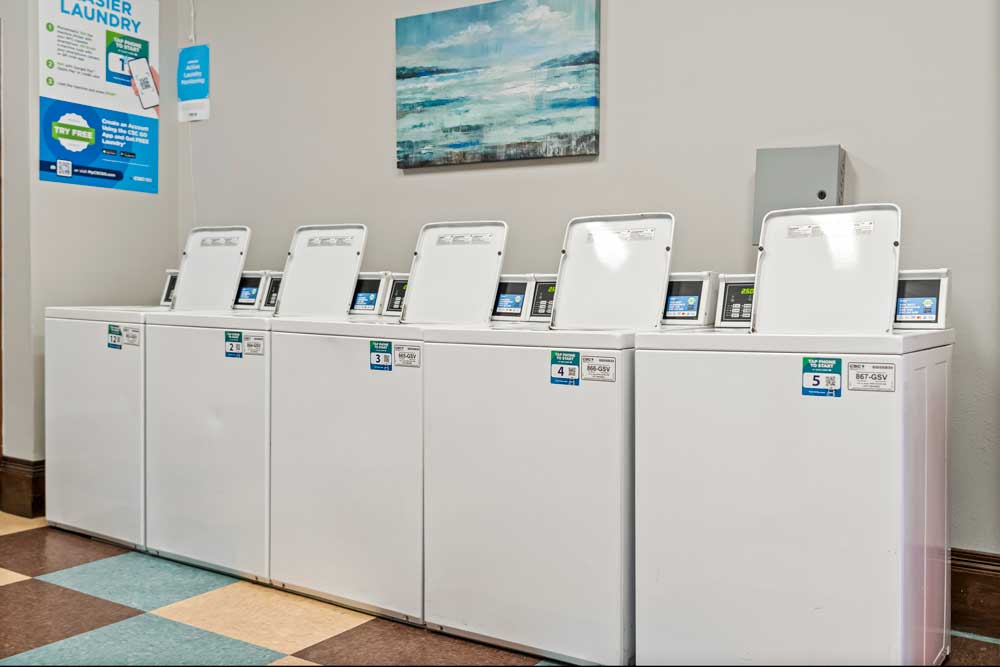 Community laundry room with multiple top-load washers, bright lighting, wall art, and clean tile flooring for resident use