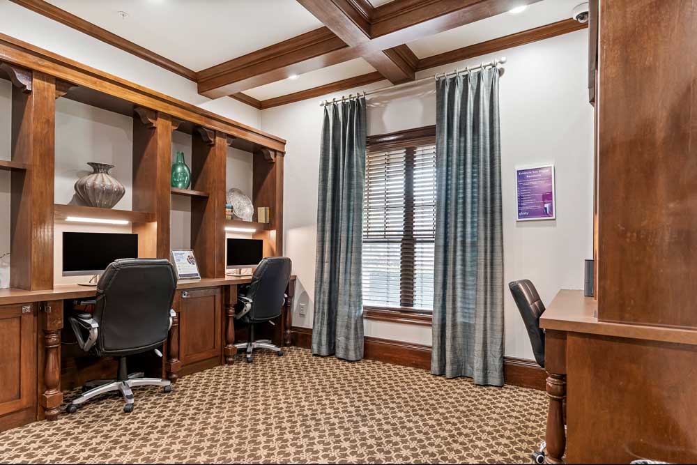 Business center with dual desktop workstations, wood cabinetry, task chairs, and window with curtains allowing natural light