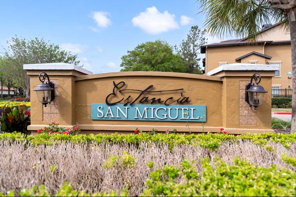 Community monument sign of Estancia San Miguel with decorative lighting, landscaped flowers, and surrounding greenery