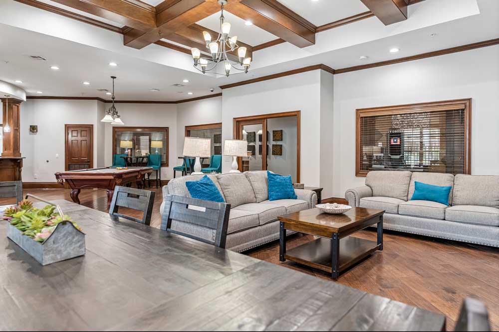 Open clubhouse lounge with sofa seating, dining table, pool table area, and large windows creating bright shared gathering space