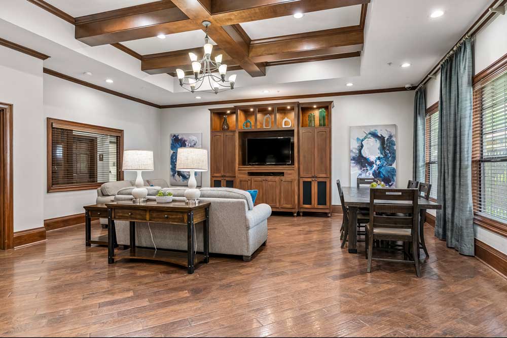 Spacious clubhouse lounge with wood beam ceilings, chandelier lighting, sofa seating, and dining area beside large windows