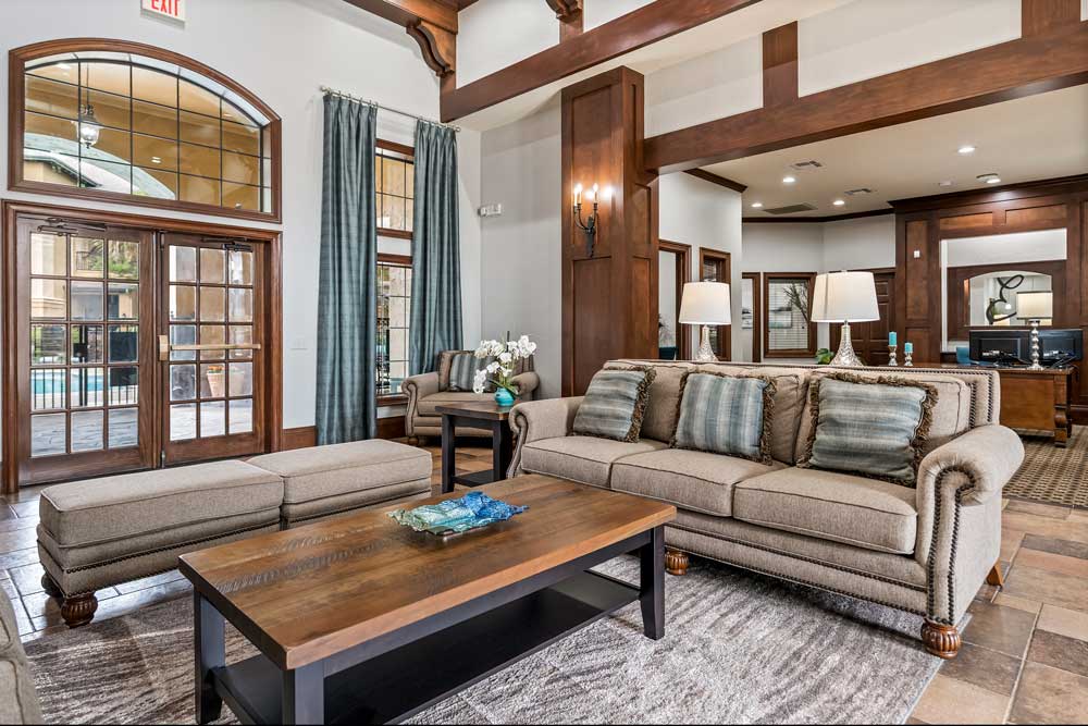 Community clubhouse lounge with sofas, ottoman seating, wood accents, large windows, and glass doors leading to outdoor pool area