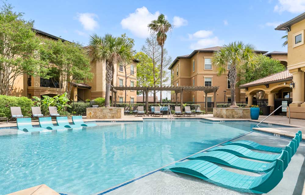 Resort-style pool with in-water loungers, pergola seating, palm trees, and apartment buildings surrounding landscaped courtyard