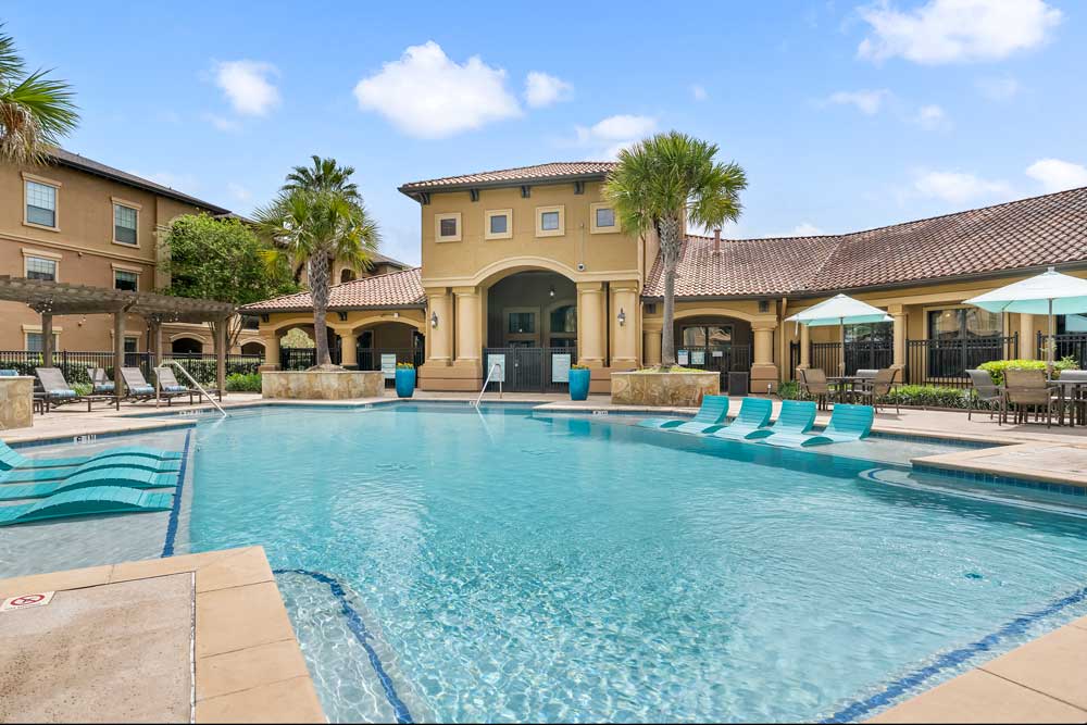 Large swimming pool with in-water loungers, clubhouse centered, palm trees, and seating areas arranged around courtyard