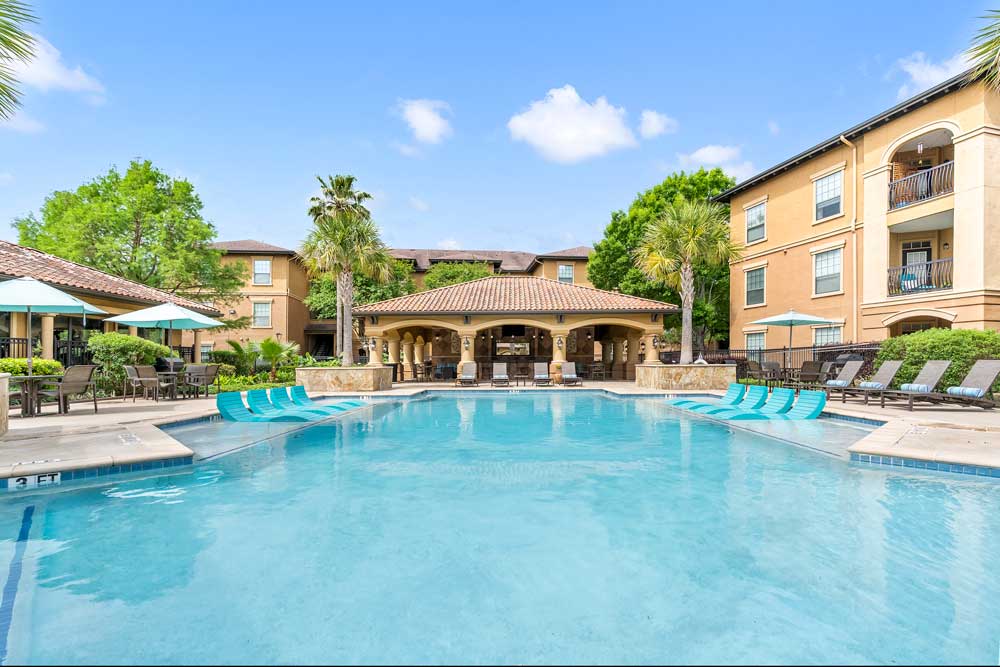 Wide pool view with in-water loungers, palm trees, umbrellas, and clubhouse centered within landscaped community