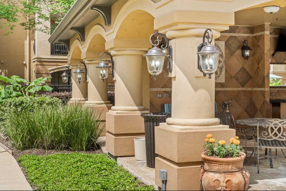 Covered patio walkway with arched columns, wall lanterns, potted plants, and landscaped greenery along apartment exterior