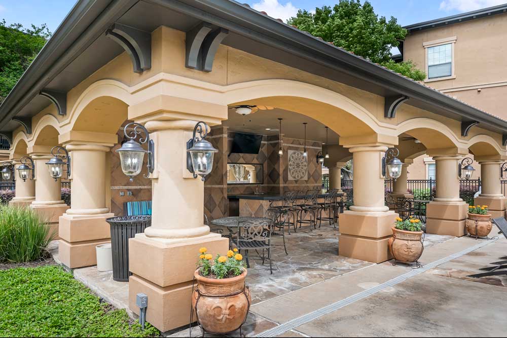 Covered outdoor kitchen and lounge with bar seating, tiled finishes, decorative lighting, and adjacent landscaped walkway