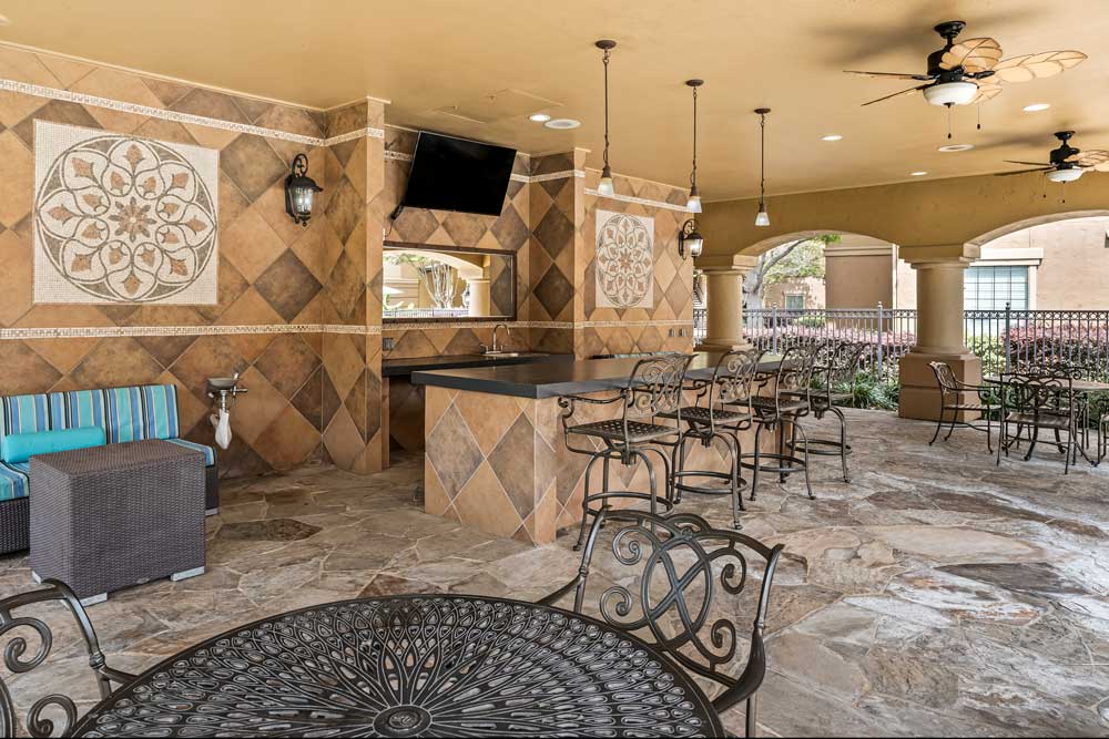 Outdoor lounge with tiled bar, seating, mounted television, pendant lighting, and decorative wall accents under covered patio