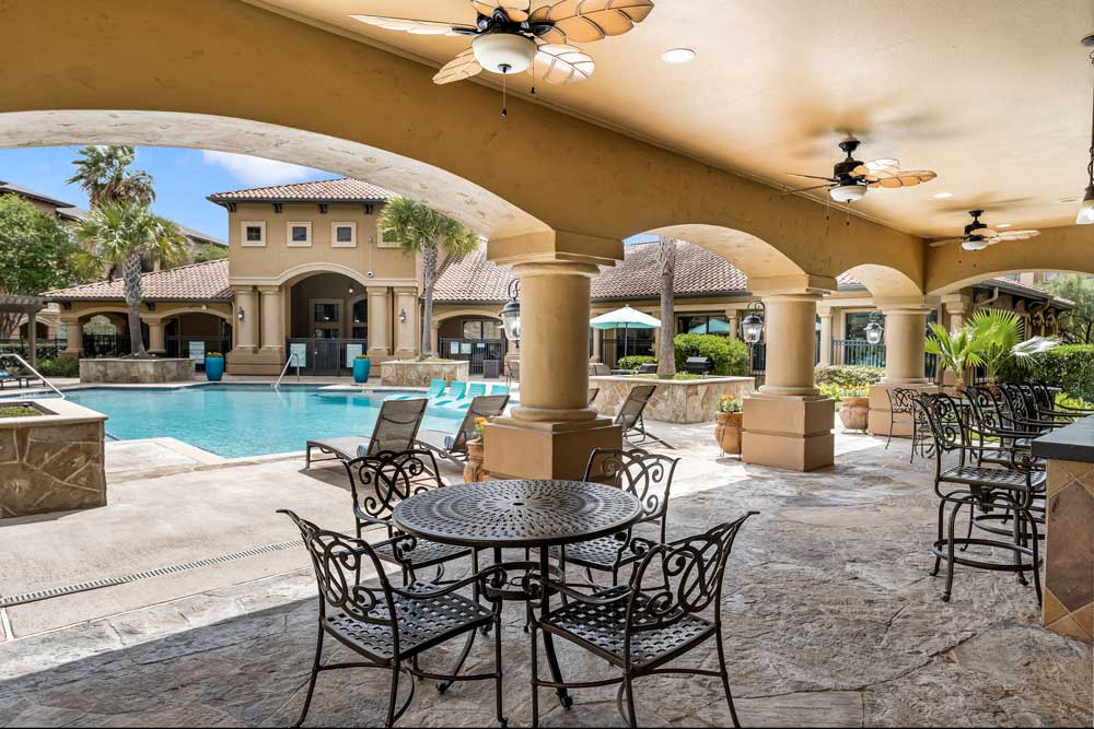 Covered poolside patio with columns, ceiling fans, seating, and view of resort-style pool and surrounding apartment buildings