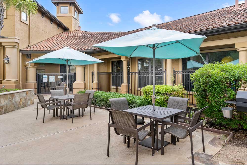 Outdoor patio with dining tables, umbrellas, and cushioned chairs set beside landscaped greenery and enclosed community pool area