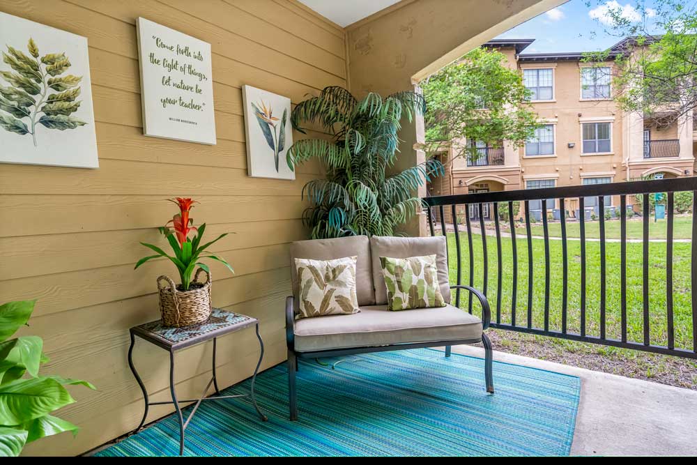 Covered patio with cushioned bench seating, decorative wall art, potted plants, outdoor rug, and railing overlooking landscaped courtyard