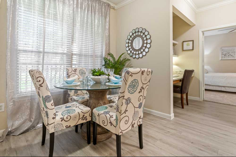 Dining area with round glass table, four upholstered chairs, large window with sheer curtains, and decorative wall mirror