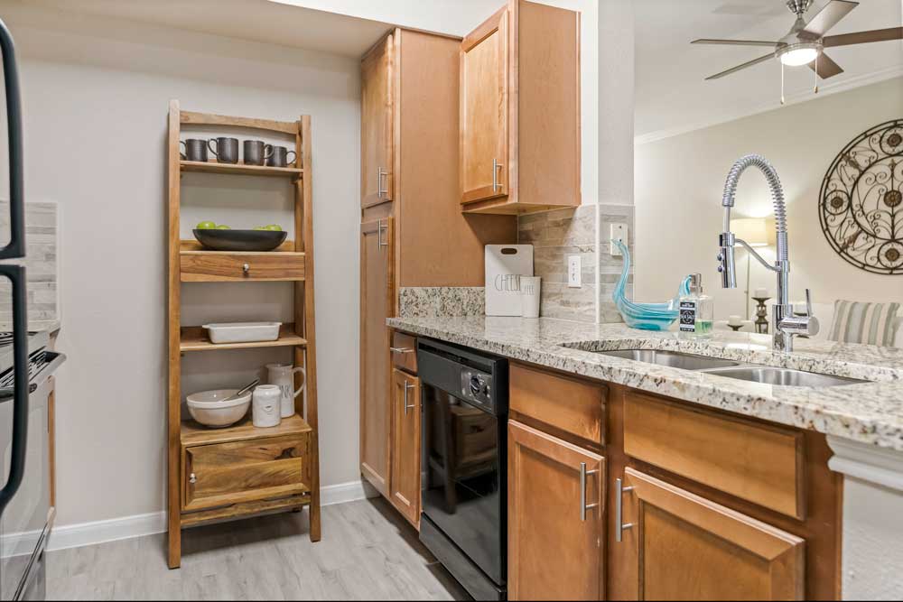 Kitchen with wood cabinetry, granite countertops, stainless sink, dishwasher, open shelving unit, and decorative accents on counter
