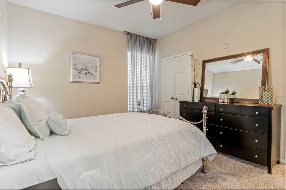 Bedroom interior featuring bed with white bedding, dresser with mirror, ceiling fan, soft carpet flooring, and window with curtains
