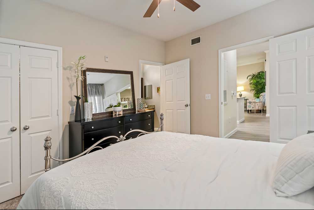 Bedroom with metal bed frame, dresser with mirror, ceiling fan, carpet flooring, and open doorway leading to kitchen and living area