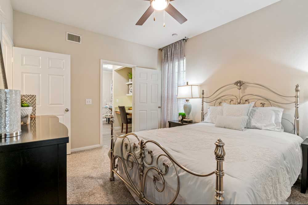 Bedroom interior with metal bed frame, nightstands, table lamps, ceiling fan, carpet flooring, and doorway leading to living area