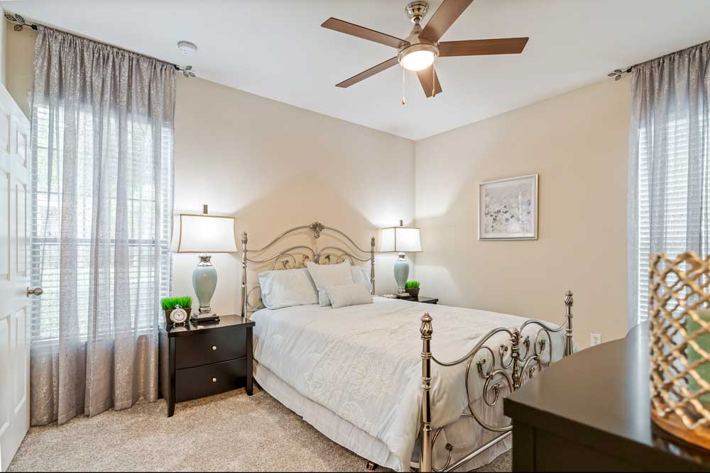 Bedroom with upholstered bed, bedside tables, table lamps, ceiling fan, soft carpeting, and large windows with curtains